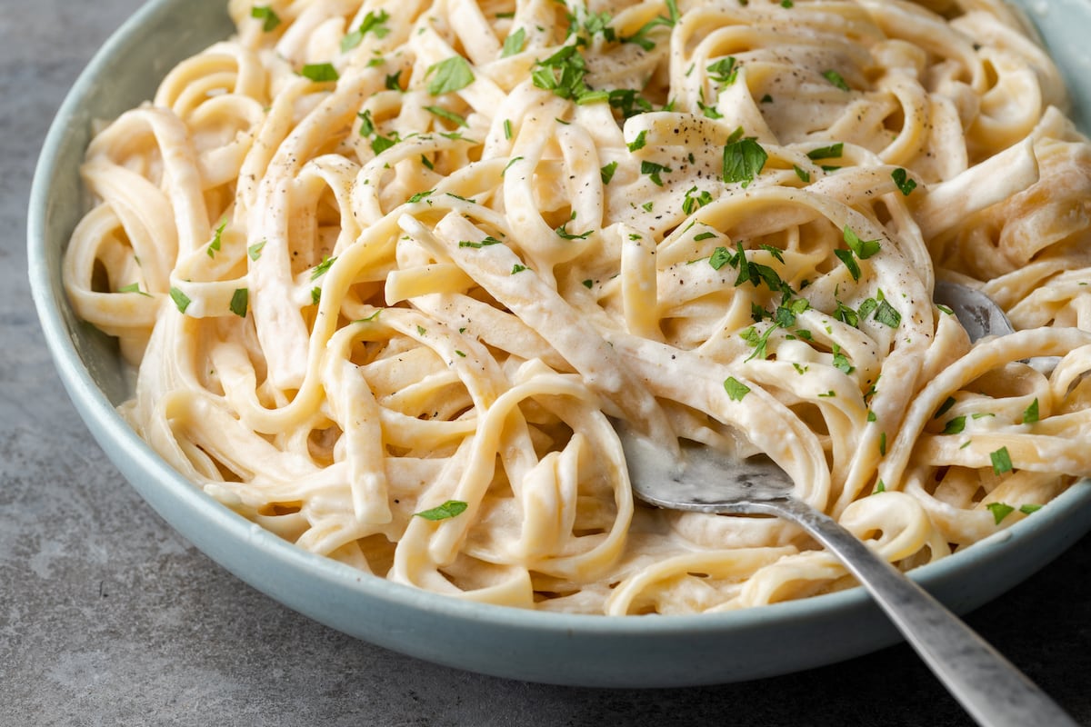 A bowl of fettuccine Alfredo garnished with fresh chopped parsley, with a silver serving utensil.